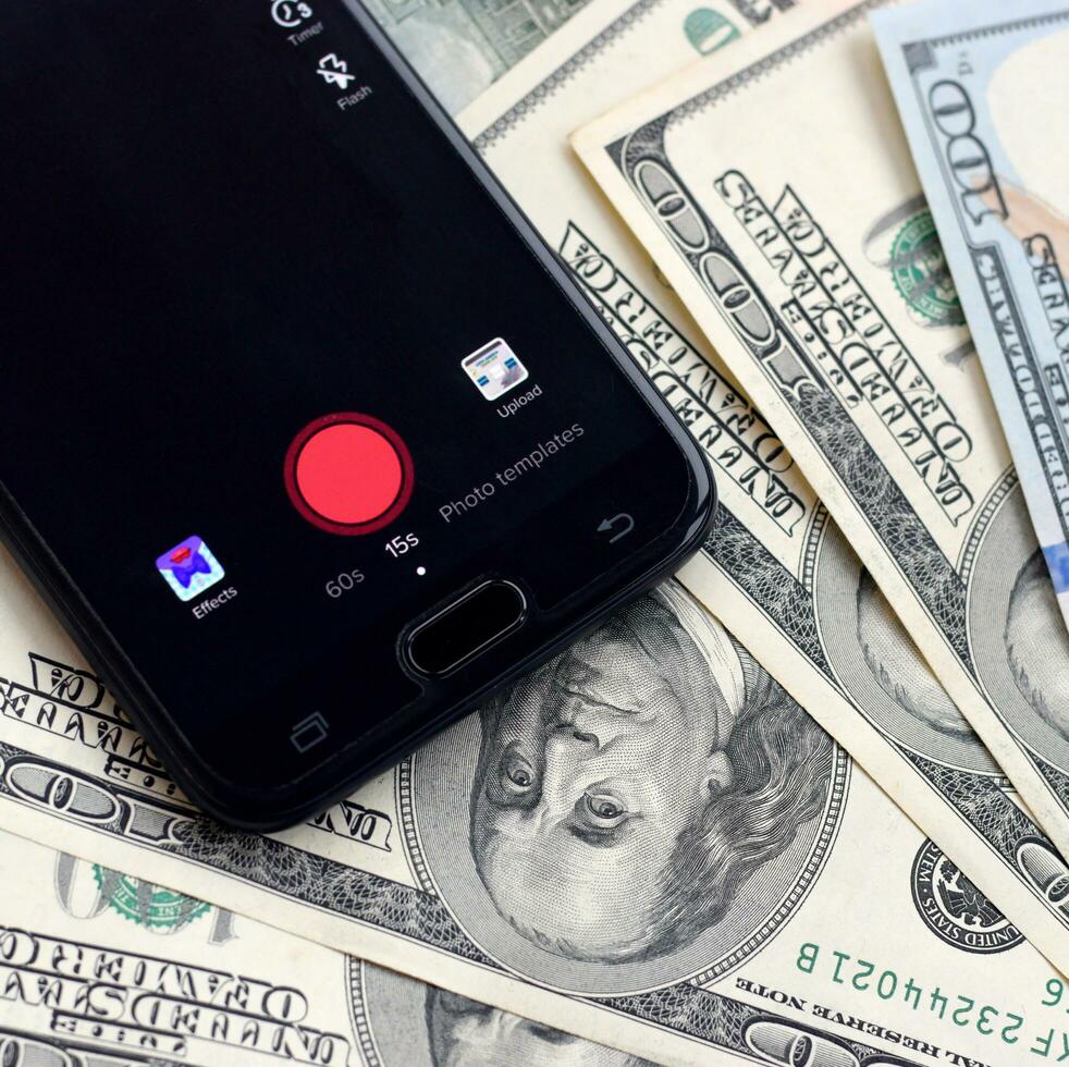 Smartphone screen with Tiktok app and lot of hundred dollar bills. Business and social networking concept photo