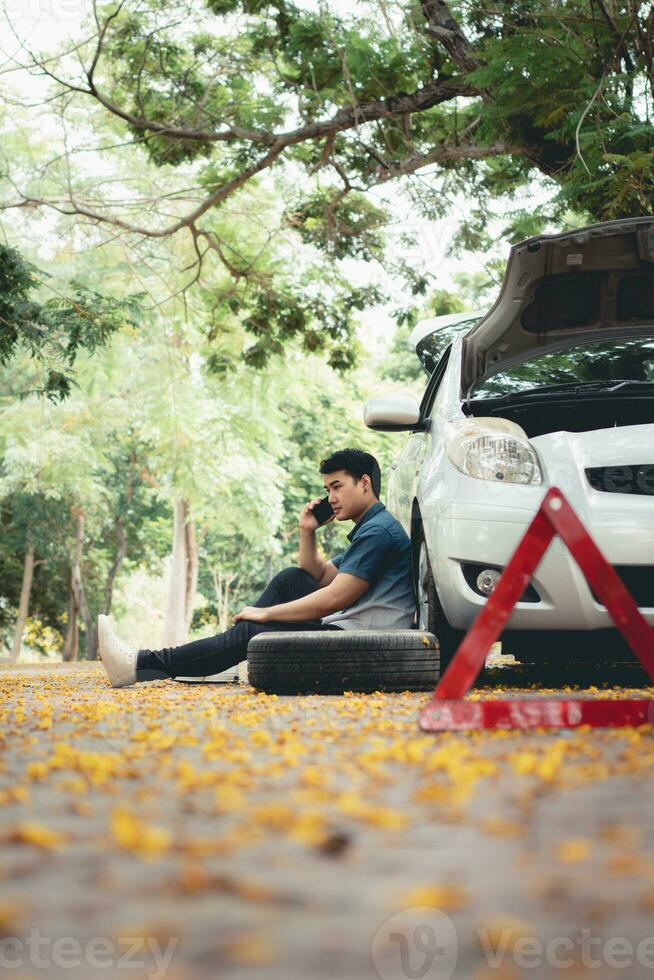 Asian man sitting beside car and using mobile phone calling for assistance after a car breakdown on street. Concept of vehicle engine problem or accident and emergency help from Professional mechanic photo