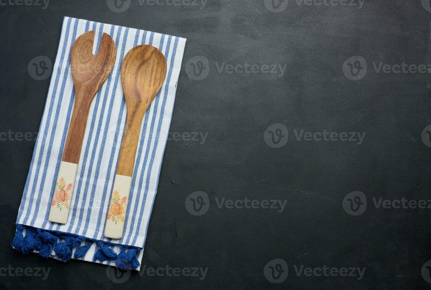 Two wooden spoons on a striped white textile kitchen towel on a black background, top view photo