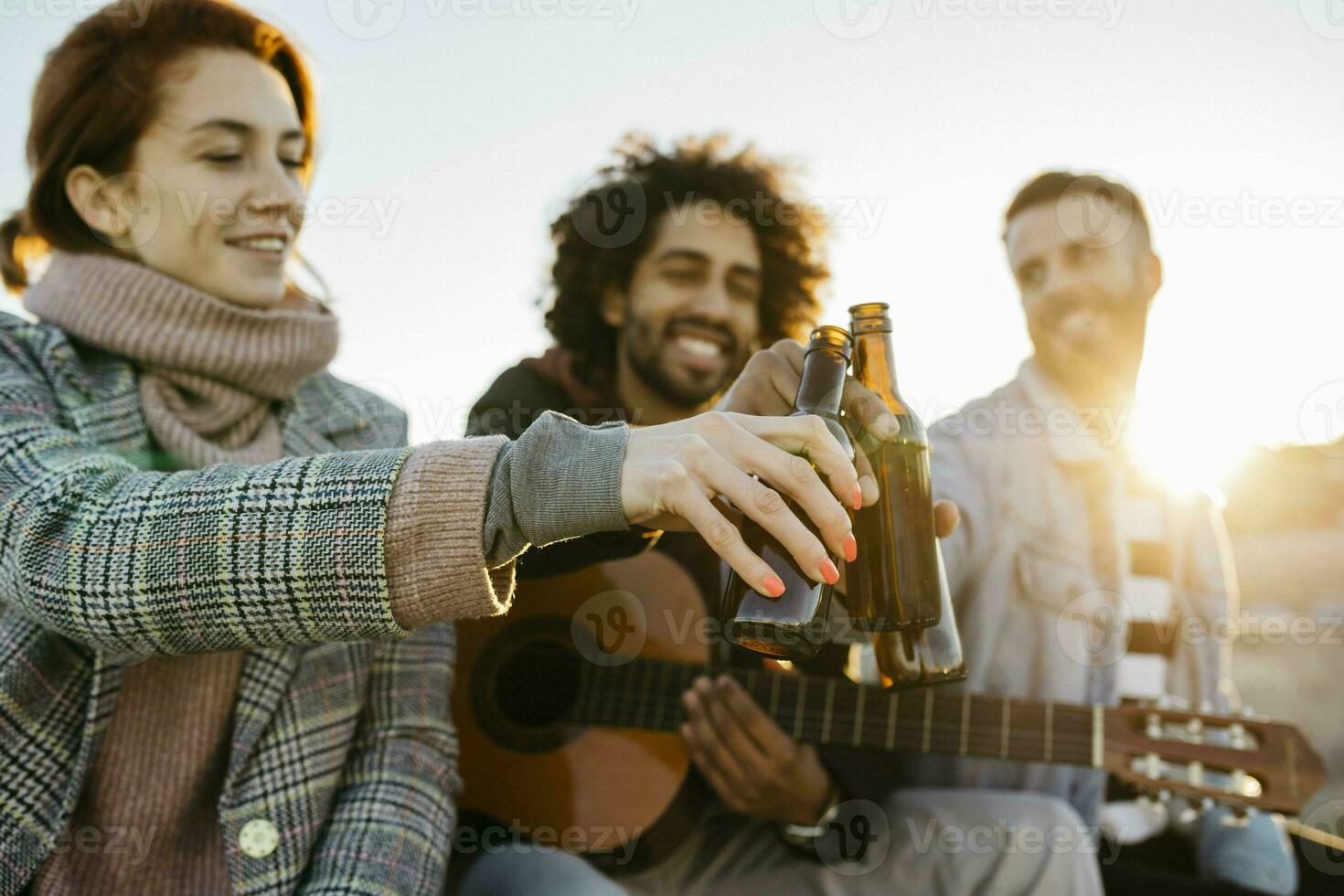 Three happy friends with guitar toasting beer bottles at sunset photo