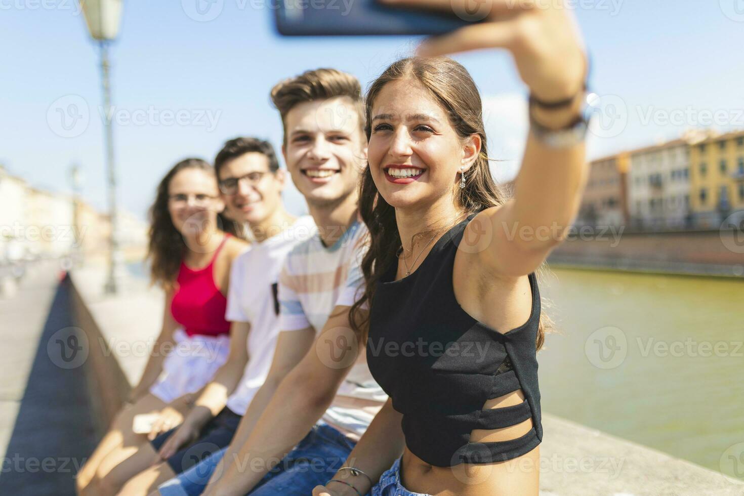 Italy, Pisa, group of four happy friends sitting on a wall along Arno river taking a selfie photo