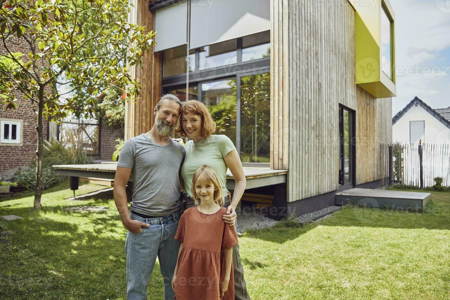 sonriente hija en pie con padres en contra minúsculo casa en yarda foto