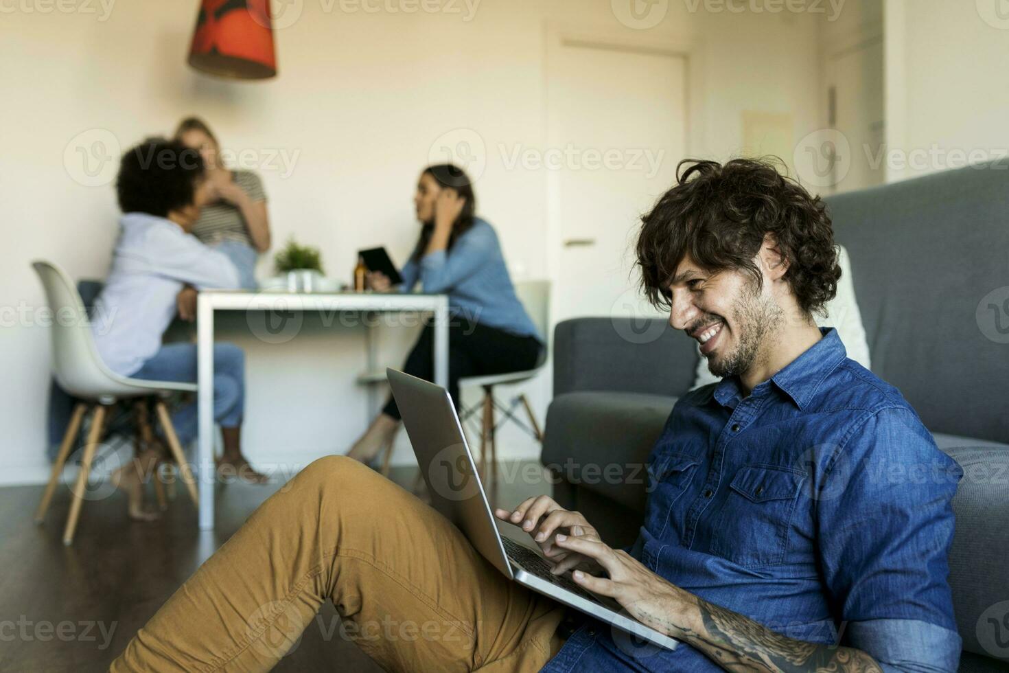 Smiling man sitting on floor using laptop with friends in background photo