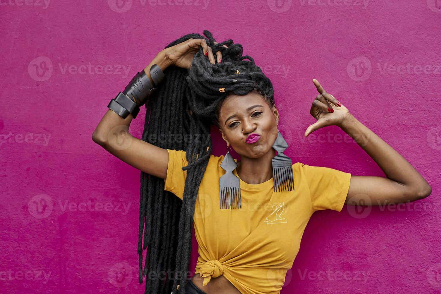 Portrait of woman with long dreadlocks making shaka sign in front of a pink wall photo