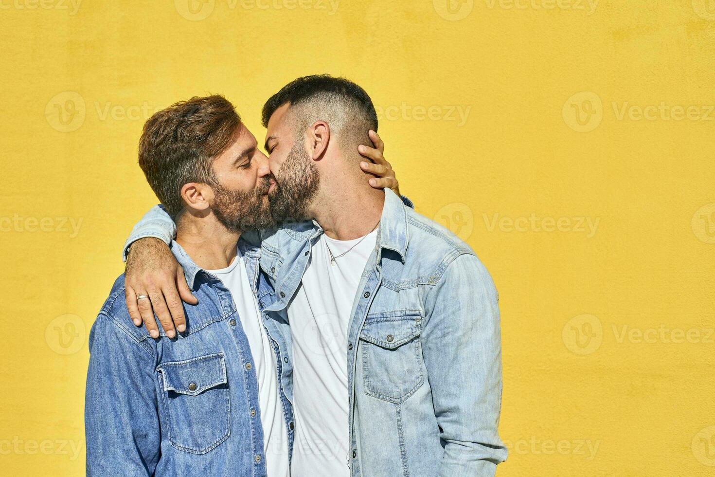 Gay couple with arm around kissing each other while standing against yellow wall photo