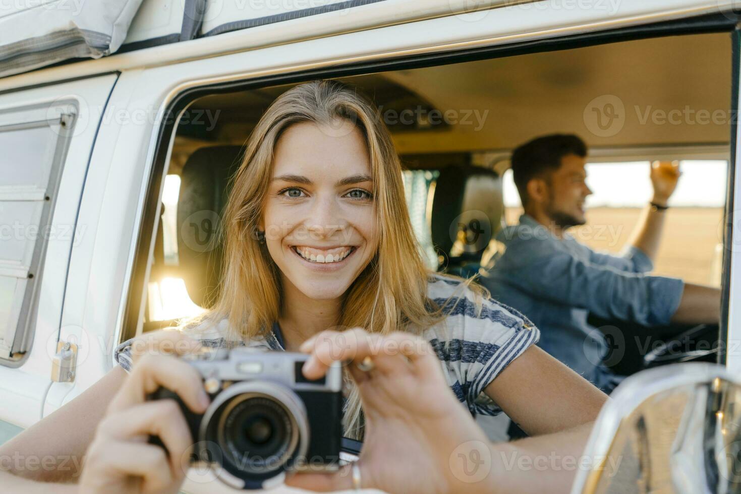 Portrait of happy woman with camera leaning out of window of a camper van with man driving photo