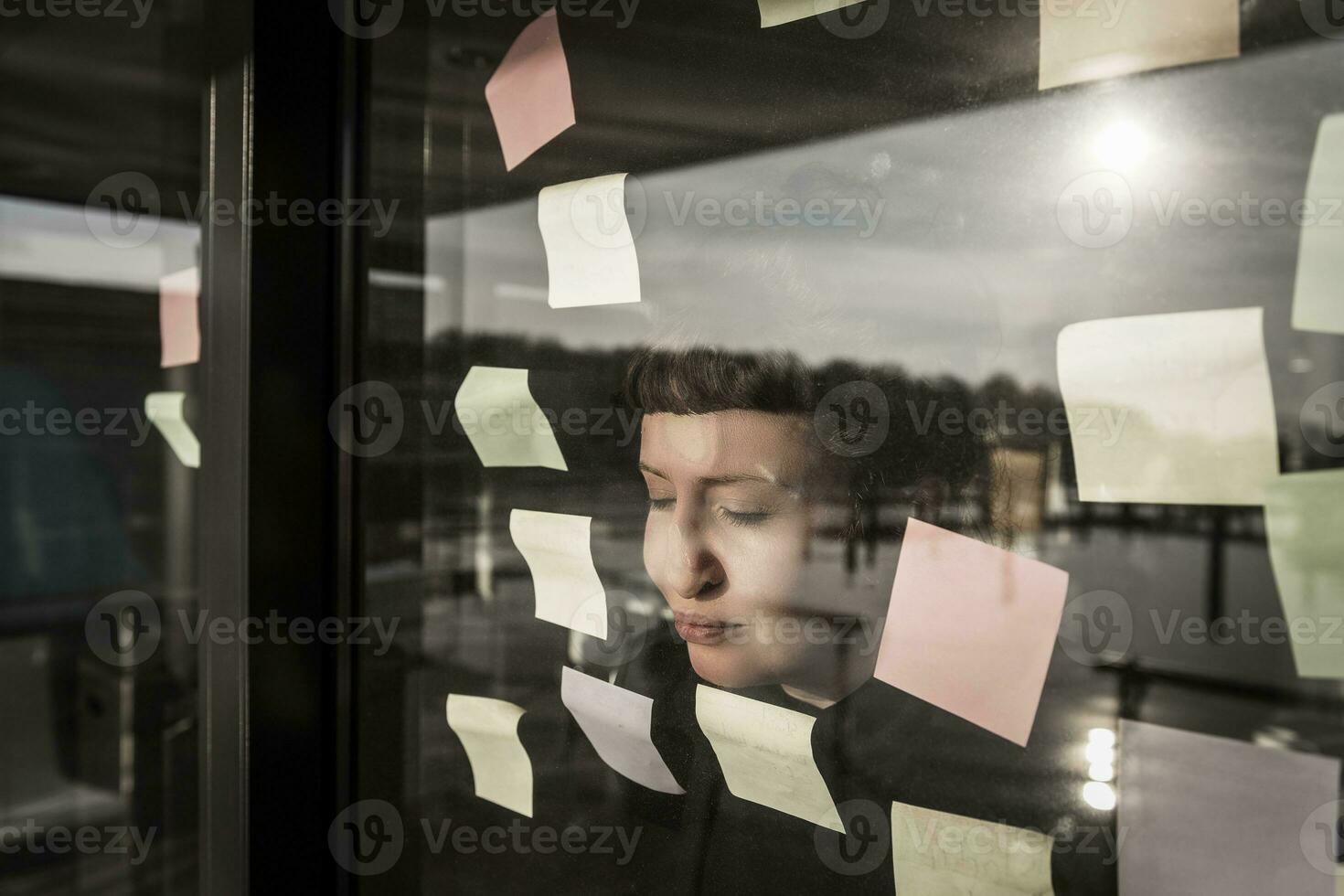 Tired woman leaning against window pane with sticky notes photo