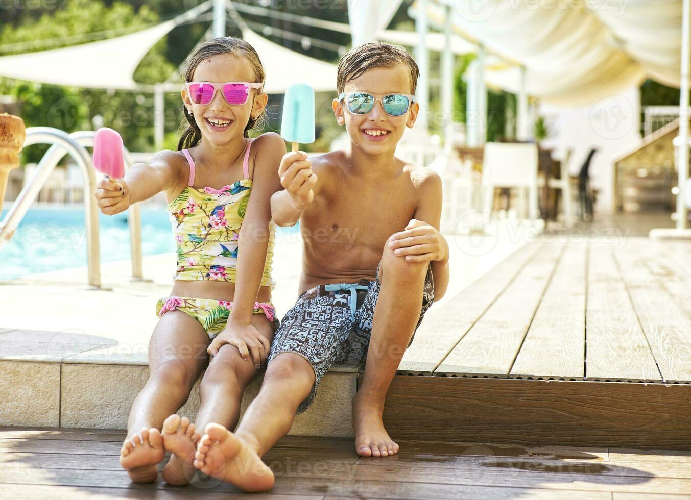 retrato de contento pequeño niña y chico vistiendo reflejado Gafas de sol demostración su Paletas de hielo foto