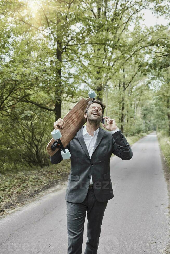 Happy businessman with skateboard talking on smartphone on rural road photo