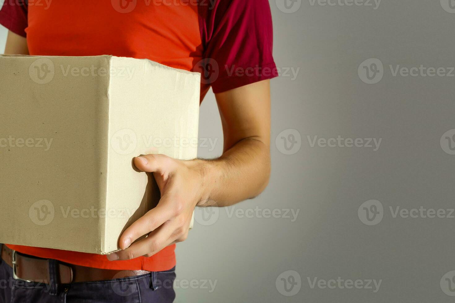 Closeup hands of delivery man holding package to deliver. Courier hand holding brown box isolated on grey background. Detail of delivery man carrying cardboard parcel with label with copy space. photo