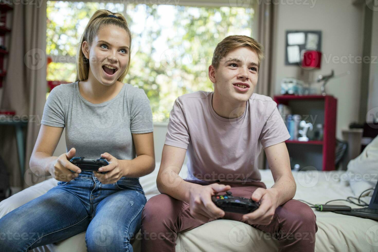 Happy teenage girl and boy sitting on bed playing video game photo