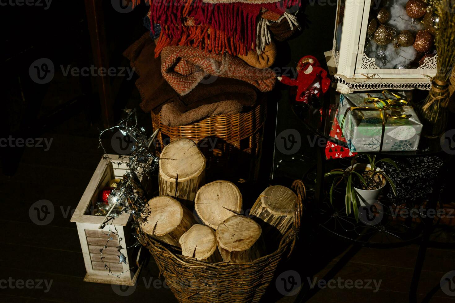 Wintertime in rustic interior. Logs for fireplace, set of cosy blankets, wooden box with Christmas baubles. Christmas garland and wrapped present. Cozy scene, dark background photo