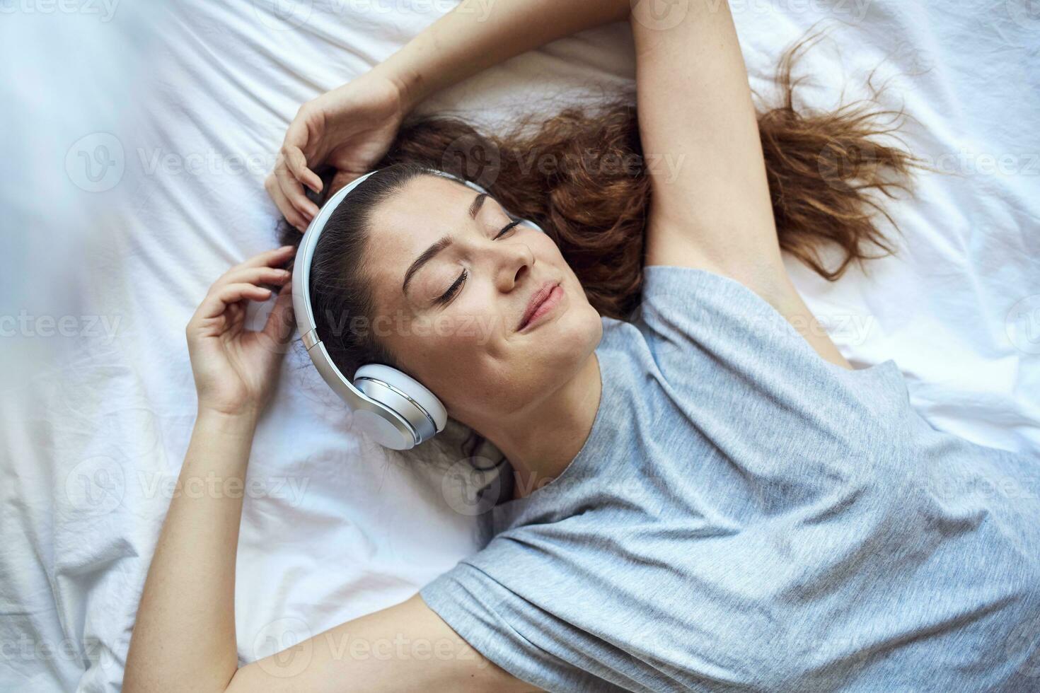 retrato de contento joven mujer con ojos cerrado acostado en cama escuchando música con auriculares foto