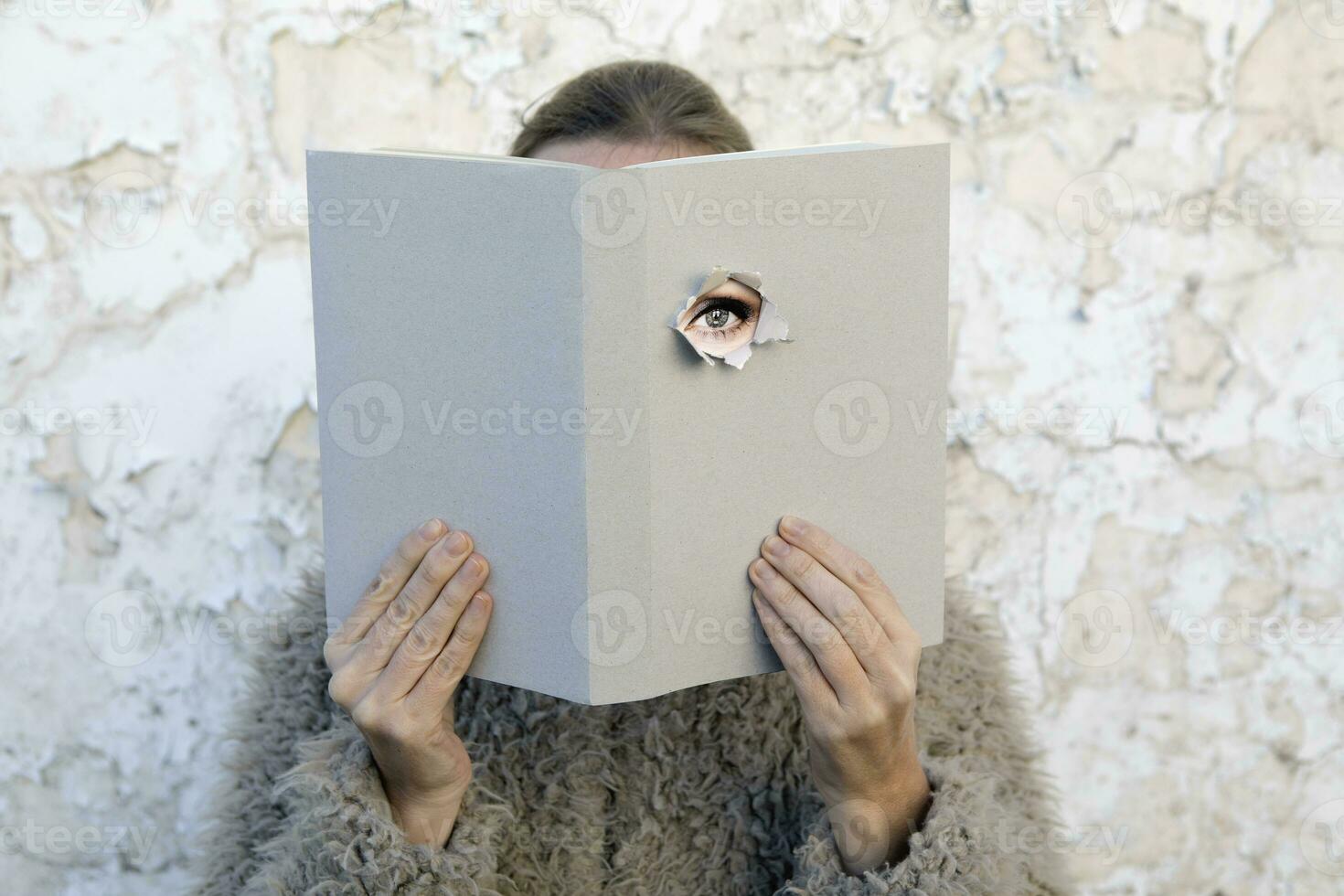 Woman covering face with book, reading poetry, eye looking through cover photo