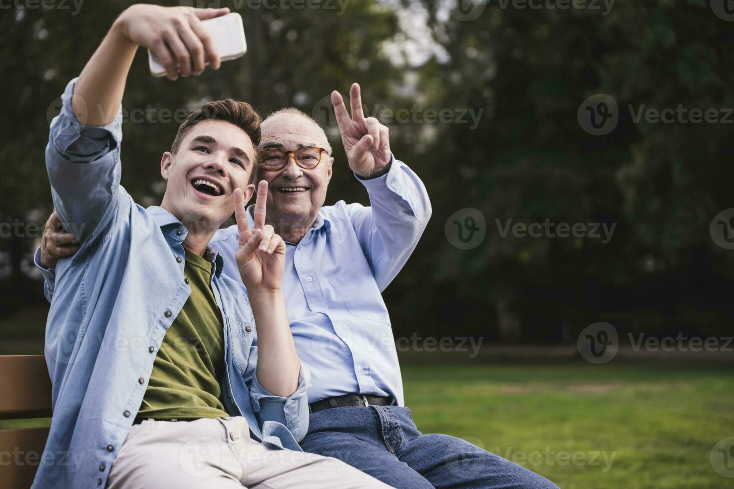 Senior man and grandson sitting together on a park bench taking selfie with smartphone photo