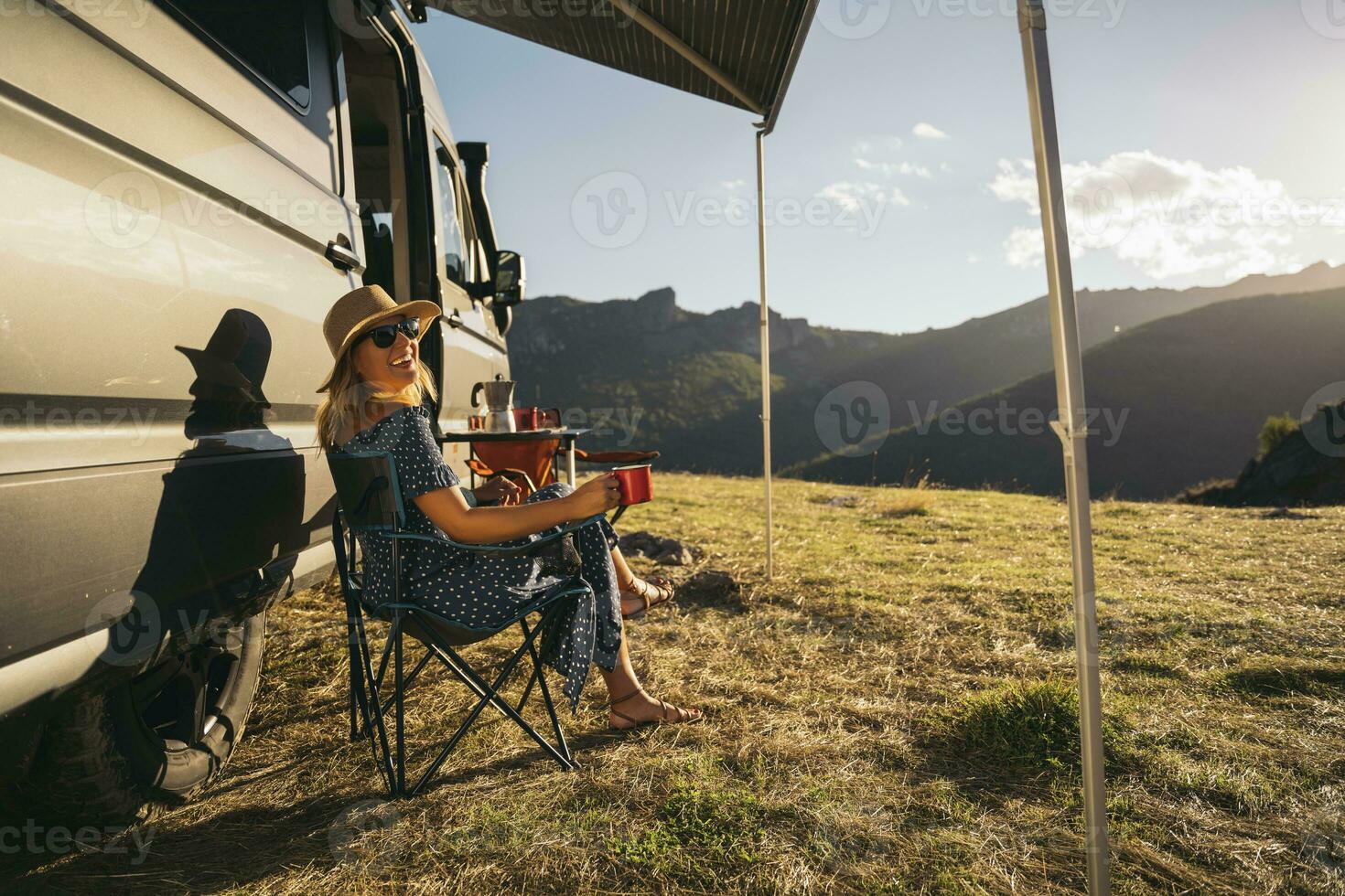 Happy woman in sunglasses with coffee cup laughing while sitting by motor home during vacations photo