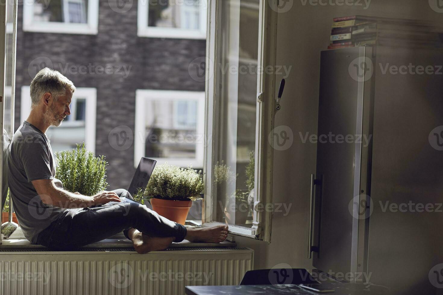 Mature man sitting at the window at home using laptop photo