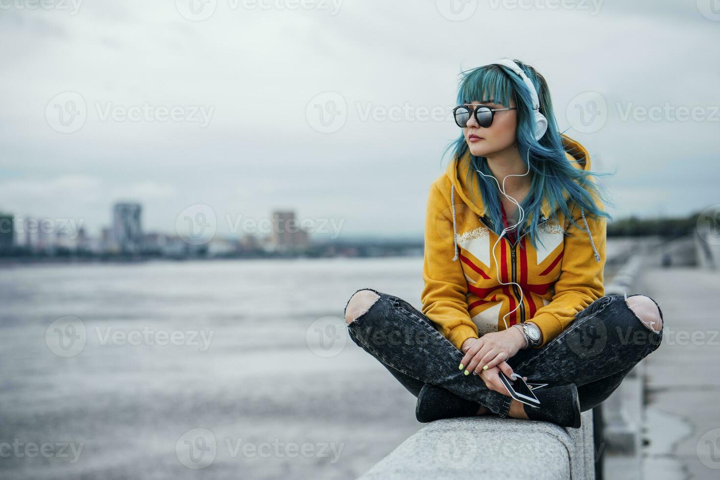 Young woman with dyed blue hair sitting on a wall listening music with headphones and smartphone photo