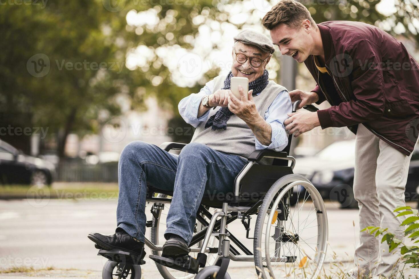 Laughing senior man in wheelchair and his adult grandson looking together at smartphone having fun photo