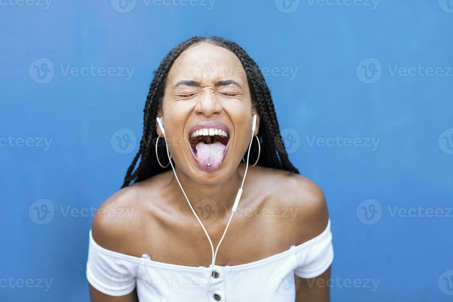 Woman listening music while sticking tongue out against blue wall photo