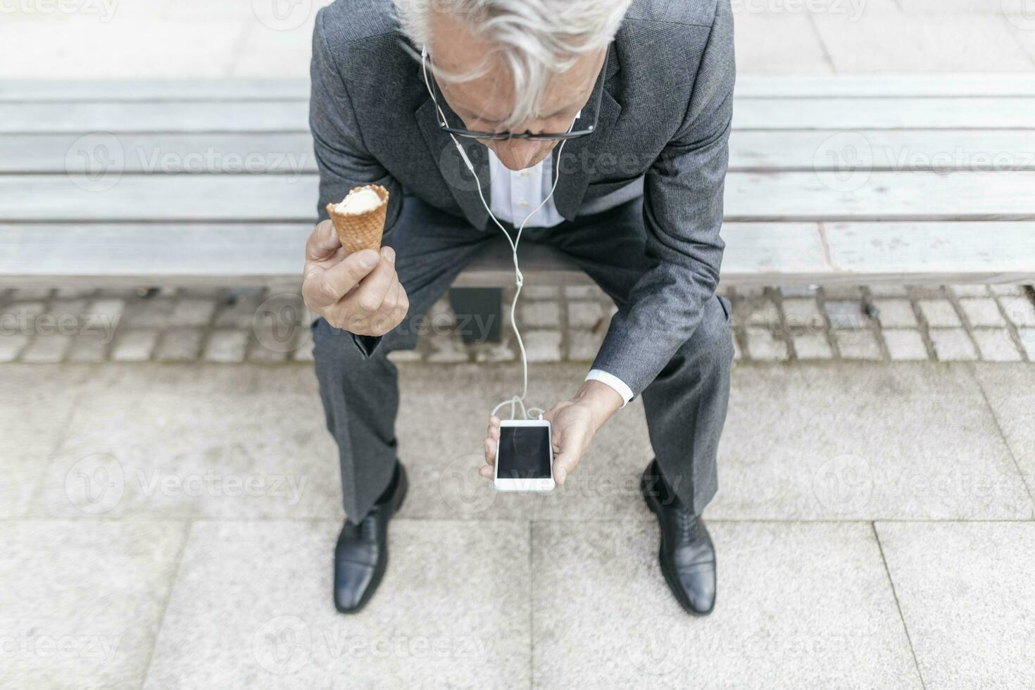 Senior businessman with ice cream cone sitting on bench looking at cell phone photo