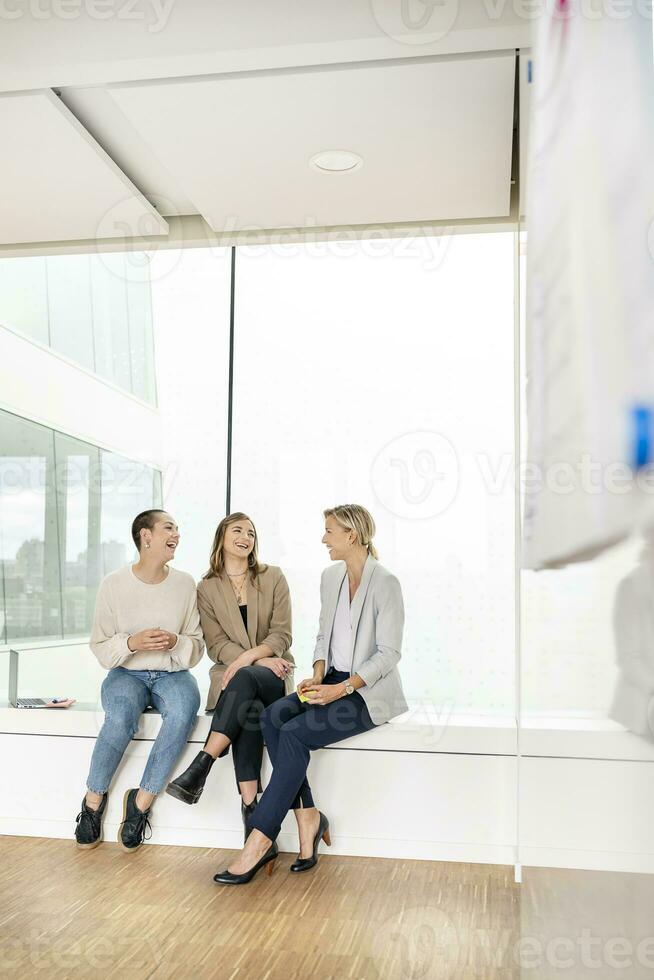 Businesswomen sitting on windowsill in office building, taking a break, talking photo