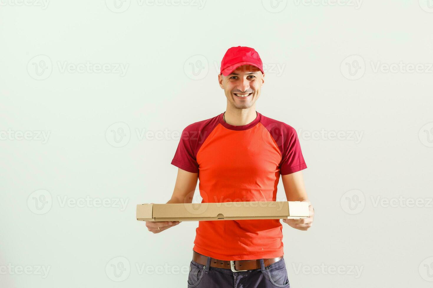 Photo of happy man from delivery service in red t-shirt and cap giving food order and holding boxes isolated over white background