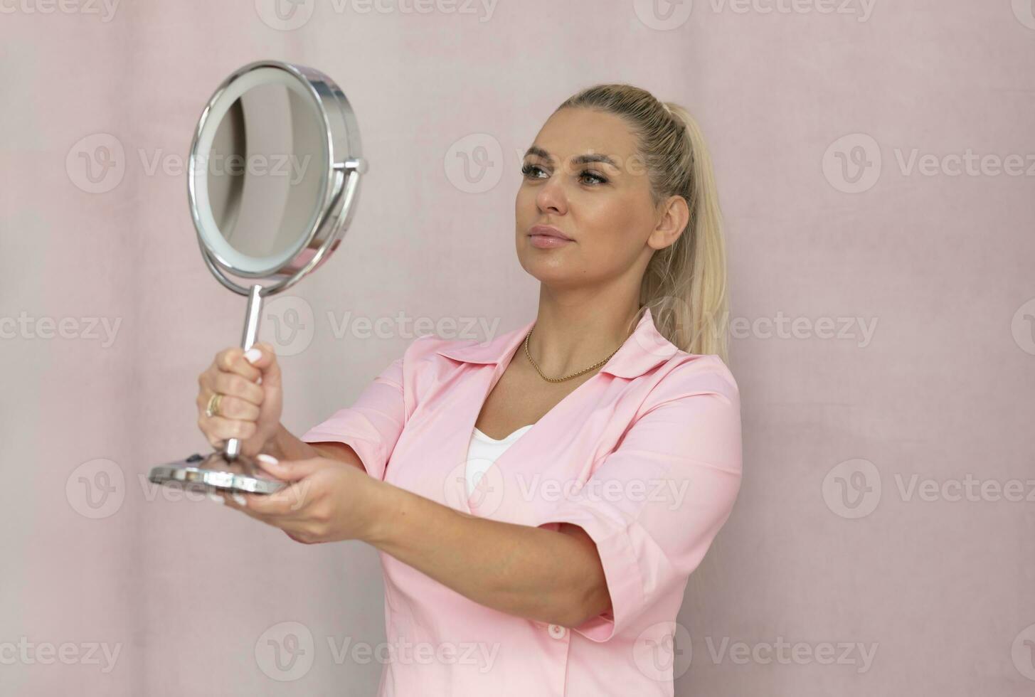 Portrait Of Attractive Real Blond Tanned Woman In Pink Dress Looking At Mirror, Fluffy Eyelashes Plump Lips On Tea Rose Color Background. Professional Caucasian Beautician Looks At Own Reflection photo