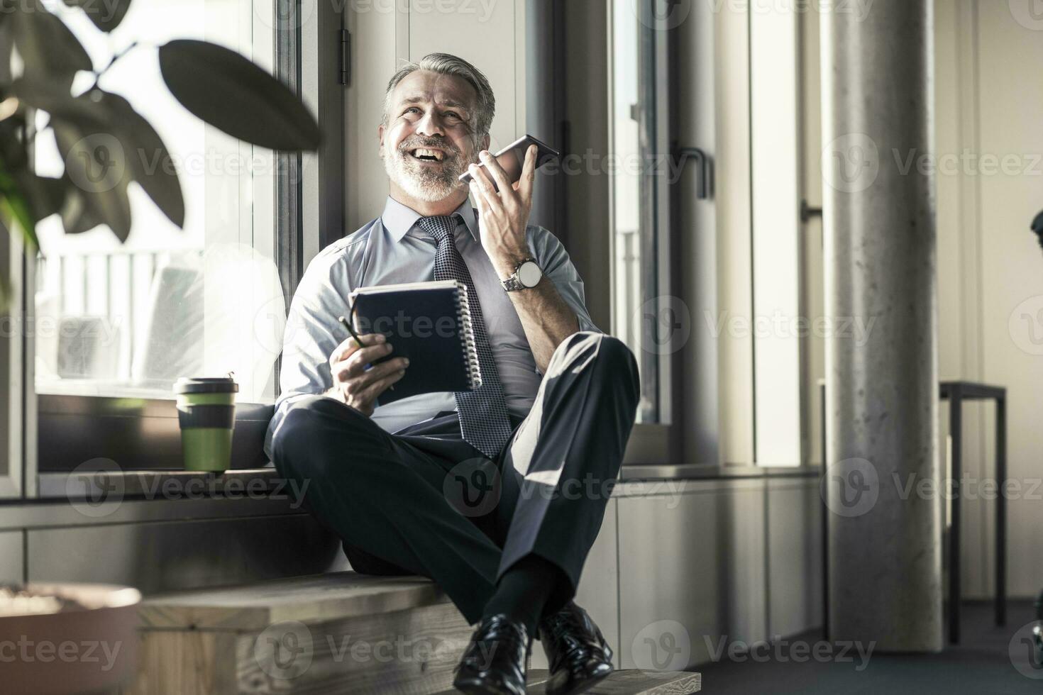 Happy mature businessman with notebook sitting at the window using cell phone photo