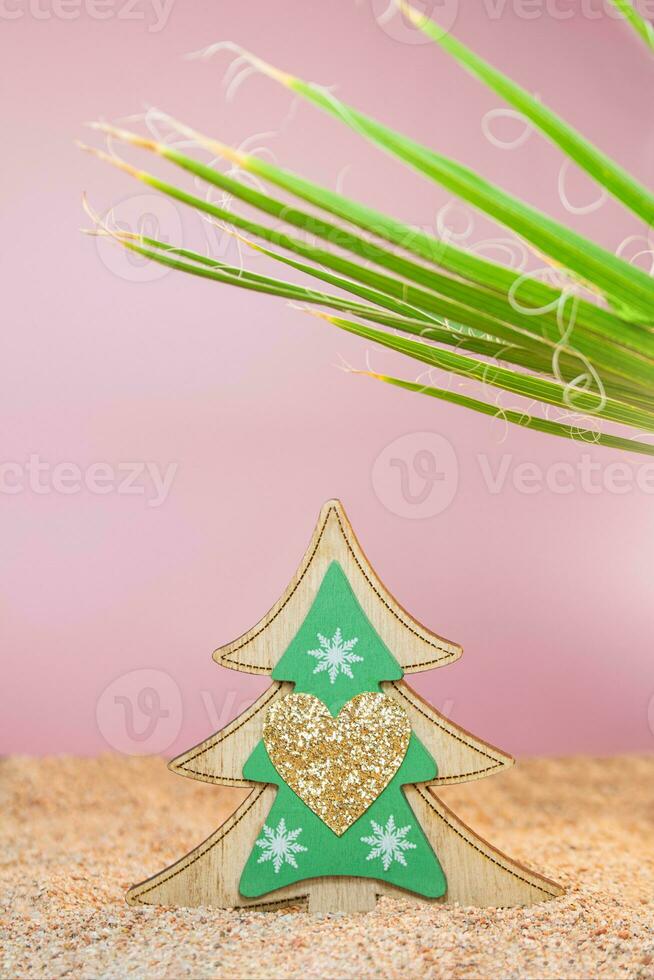 Wooden Christmas tree with sparkles in the sand with a palm tree on a pink background. Holidays in hot countries. Vertical. Copy space photo