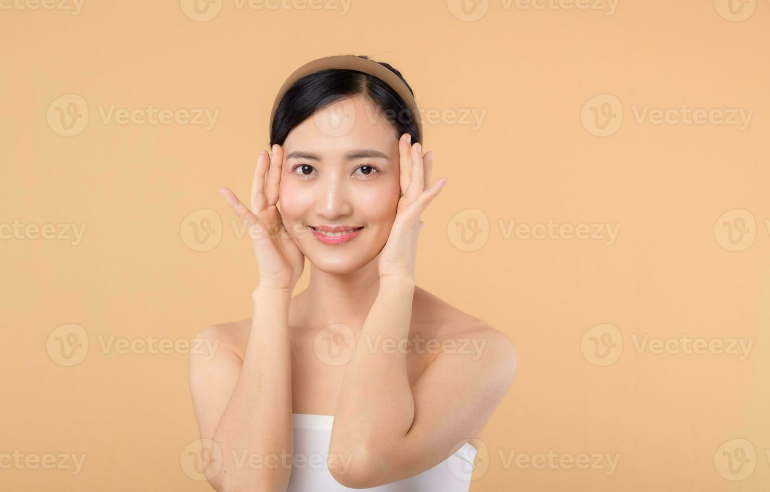 Beautiful girl asian model touching fresh glowing hydrated facial skin on beige background closeup. Beauty face young woman with natural makeup and healthy skin portrait. Skin care concept photo