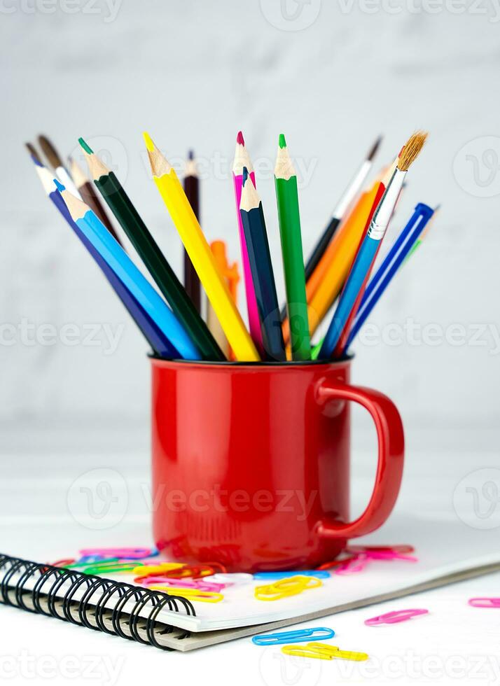 Bright pencils and brushes in a red mug on a white wooden table. School supplies. Back to school. Close-up. Selective focus. photo