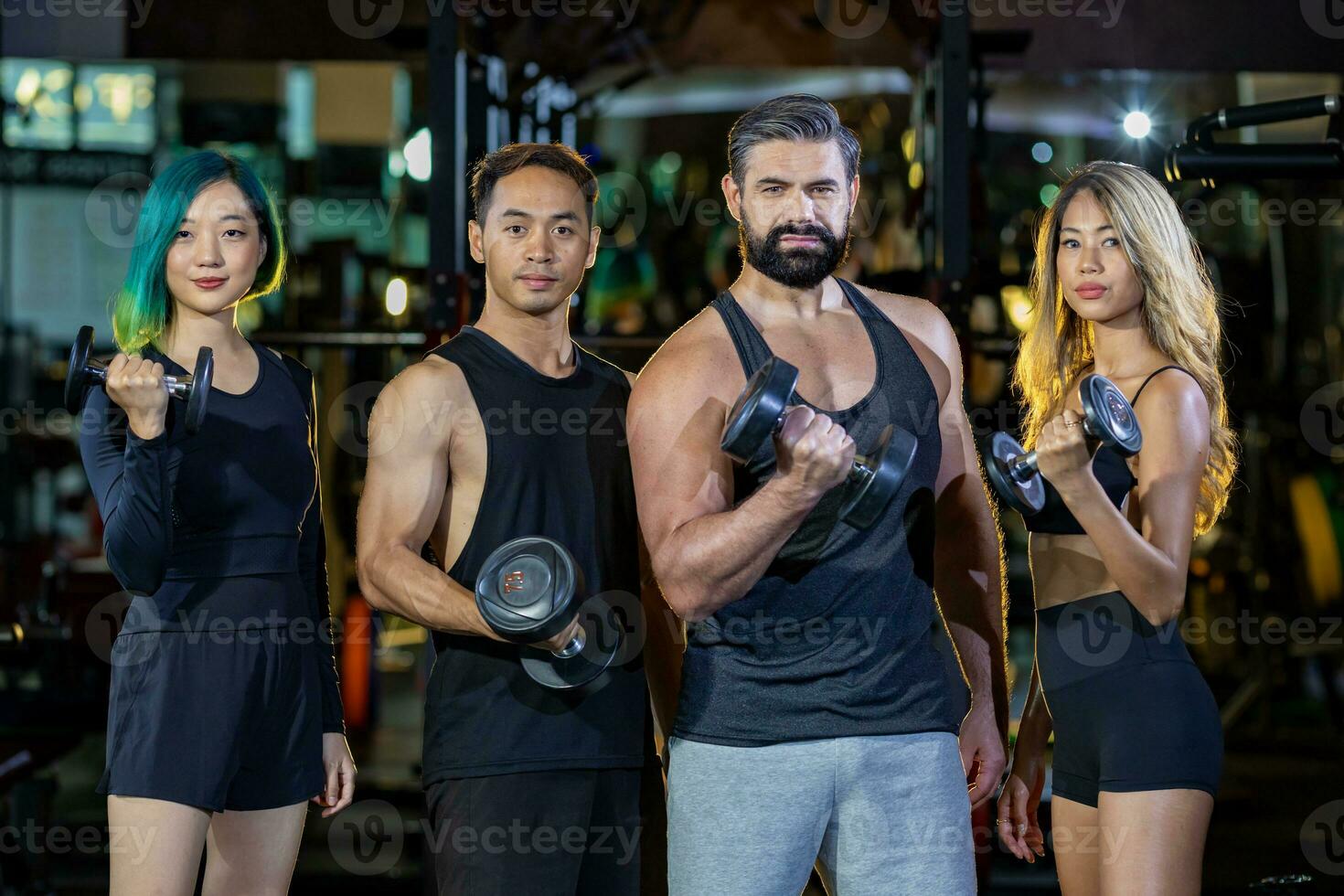 Group portrait of diversity muscular gym trainers standing together while lifting dumbbell inside gym with dark tone background for exercise and workout photo