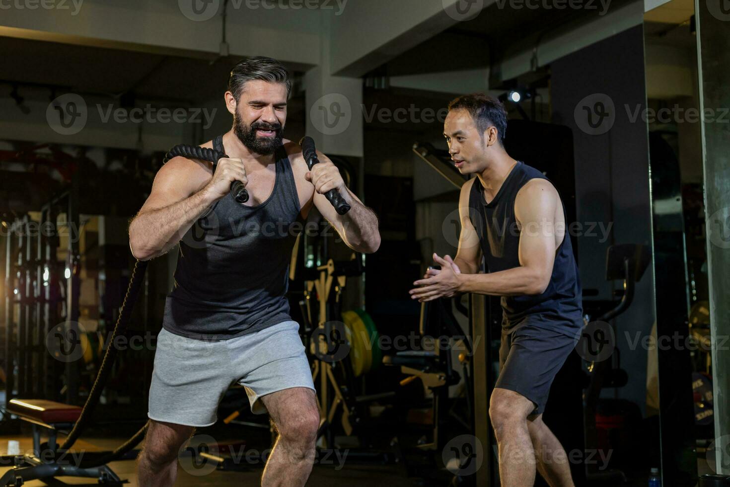 Caucasian man with strong and muscular body training on core weight training by rope pulling with his supporting trainer encouraging inside gym for advance exercise and workout concept photo