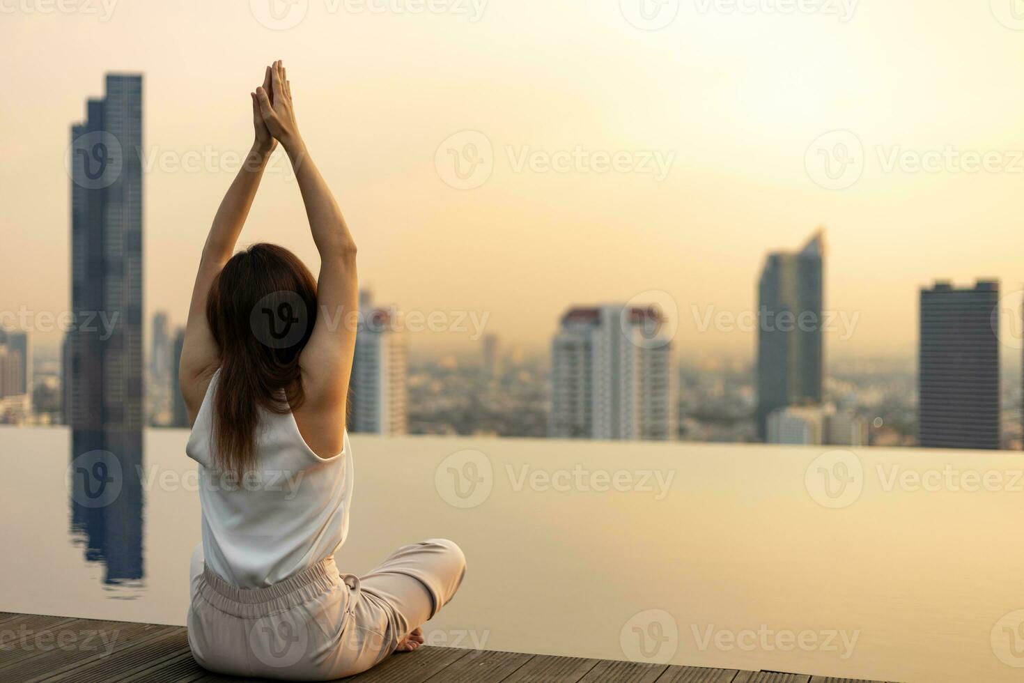 Back of woman relaxingly practicing yoga meditation at the swimming pool rooftop with view of urban skyline building to attain happiness from inner peace wisdom photo