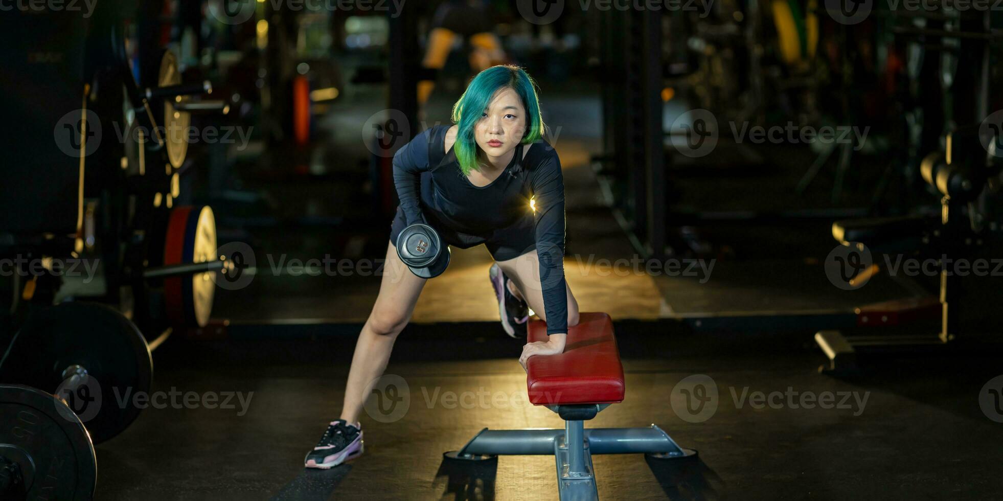 asiático mujer con fuerte y muscular cuerpo formación en doblado terminado fila pesa con banco curioso a el cámara a construir espalda y ala músculo dentro el gimnasio con oscuro tono estilo foto