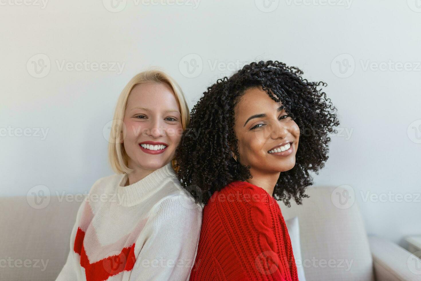 Two beautiful women , blond and african, wearing knitted jumpers sitting on the sofa, back to back and smilling, Cheerful women back against back looking at camera and sitting on the sofa photo