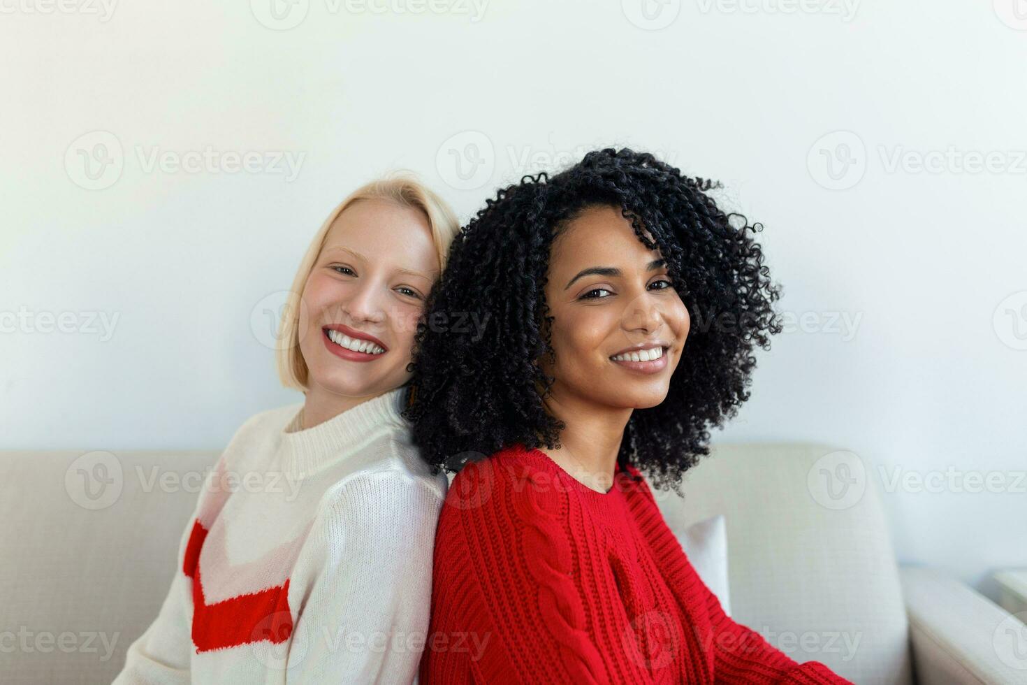 Two beautiful women , blond and african, wearing knitted jumpers sitting on the sofa, back to back and smilling, Cheerful women back against back looking at camera and sitting on the sofa photo