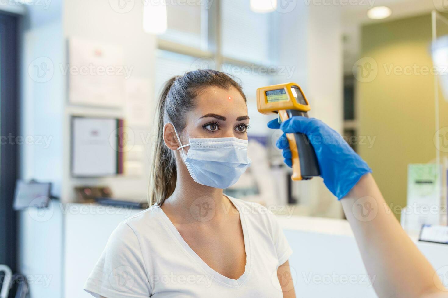 Nurse checking woman's temperature with infrared thermometer at counter. Female is wearing protective face mask. They are at hospital during pandemic. photo