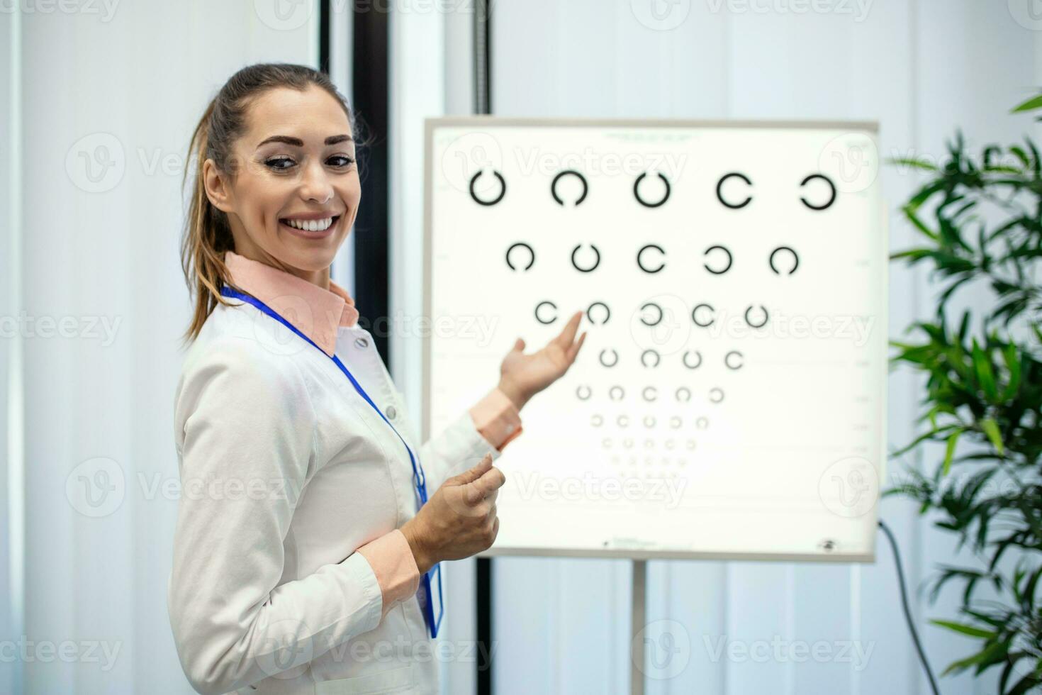 Professional female optician pointing at eye chart, timely diagnosis of vision. Portrait of optician asking patient for an eye exam test with an eye chart monitor at his clinic photo