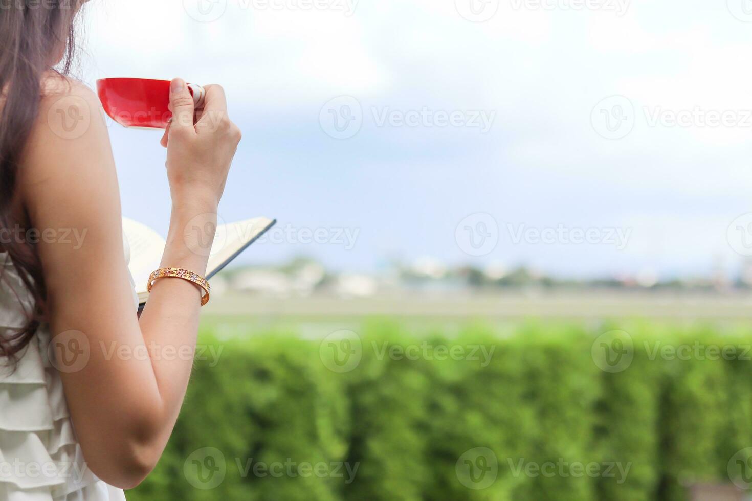 A young woman wearing a sexy white see through nightgown stands by the window with a red cup of coffee in the morning to drink coffee and see the beautiful scenery of the sunrise in the morning. photo