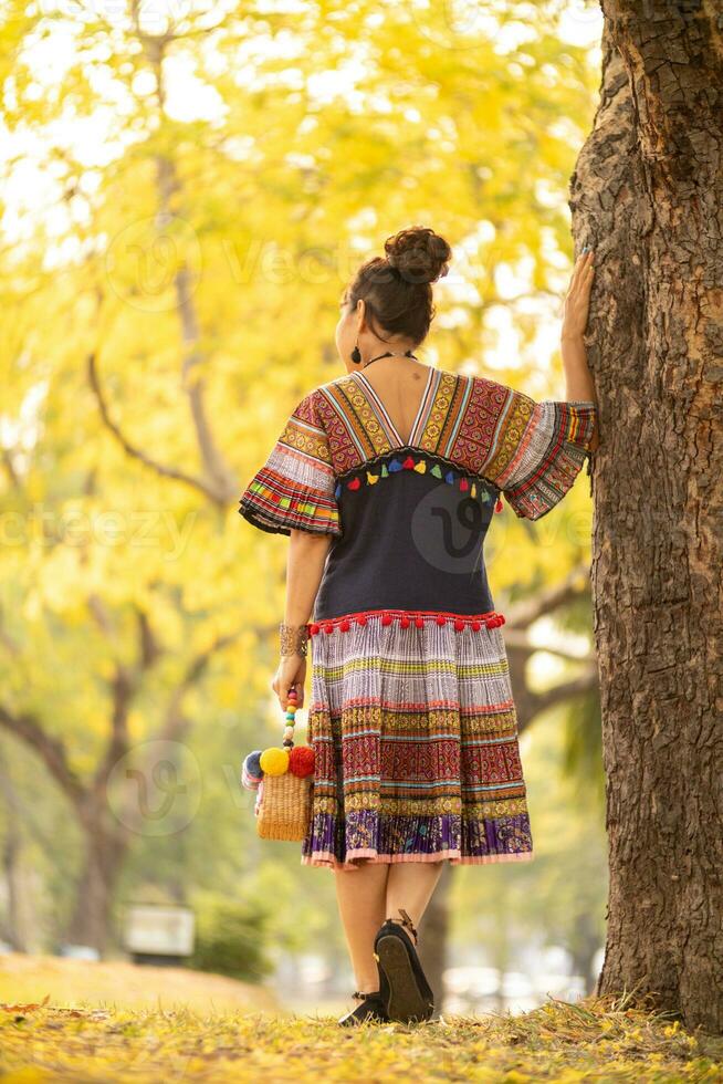 An elderly woman wearing fashionable dress using traditional tribal fabric designed to match era and standing under beautiful yellow flowering tree. An elderly woman stood alone under yellow flower. photo
