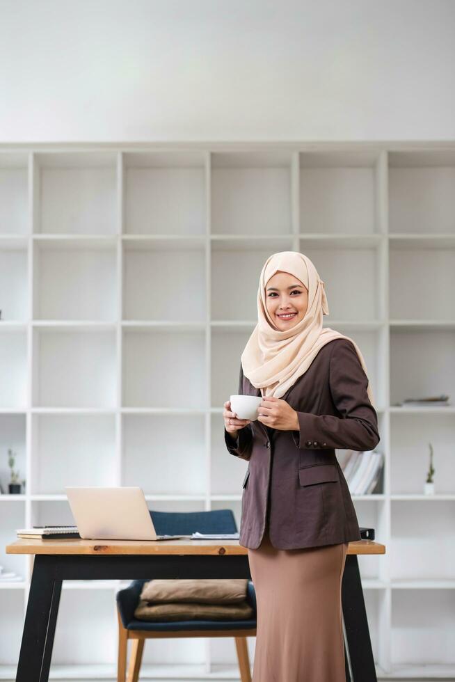 un retrato de un confidente y sonriente asiático musulmán mujer de negocios soportes en su moderno oficina con un café taza en su manos. foto