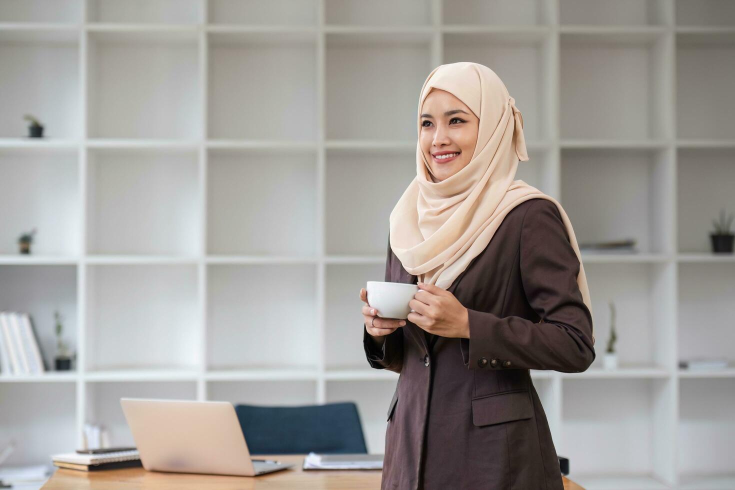 un retrato de un confidente y sonriente asiático musulmán mujer de negocios soportes en su moderno oficina con un café taza en su manos. foto