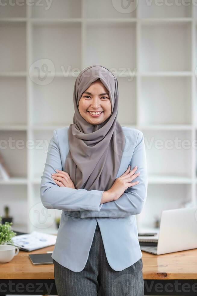 un retrato de un hermosa y profesional asiático musulmán mujer de negocios soportes en el oficina con su brazos cruzado. foto