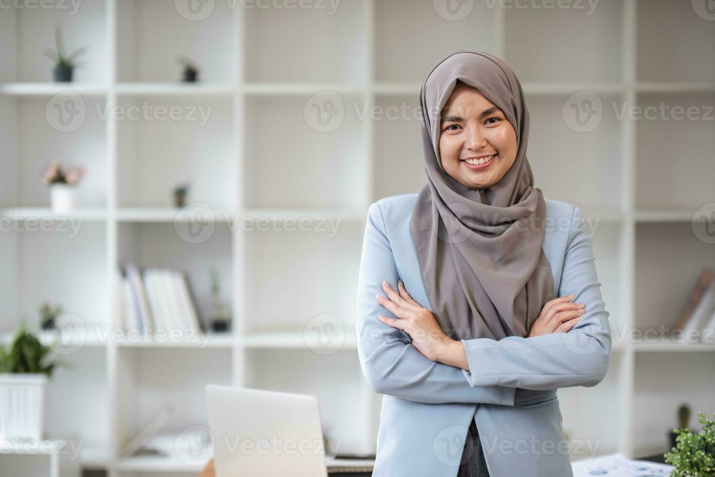 un retrato de un hermosa y profesional asiático musulmán mujer de negocios soportes en el oficina con su brazos cruzado. foto