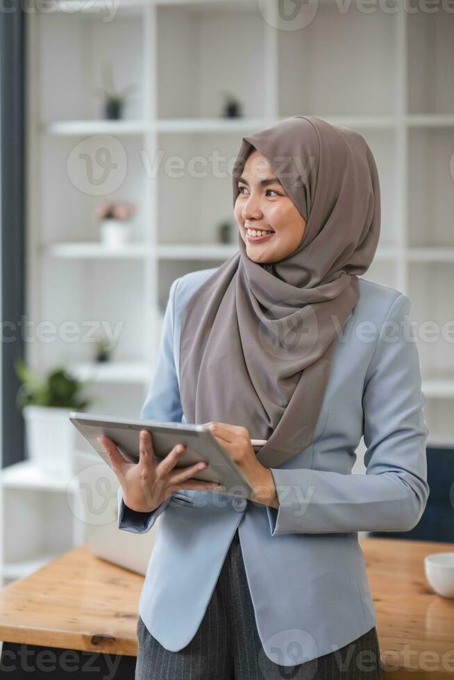 un retrato de un exitoso asiático musulmán mujer de negocios soportes en su moderno oficina con un digital tableta en su mano. foto