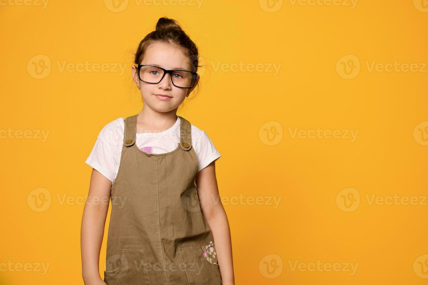 Mischievous child girl in stylish casual clothes and eyeglasses, looking at camera, isolated orange studio background photo