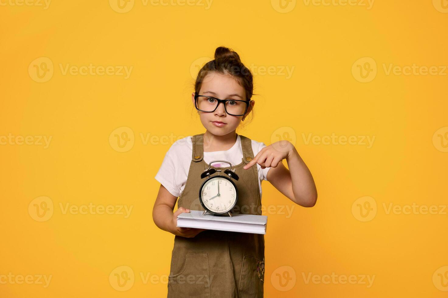 linda primario colegio niña en los anteojos, participación un libro y demostración un negro alarma reloj y mirando con confianza a cámara foto