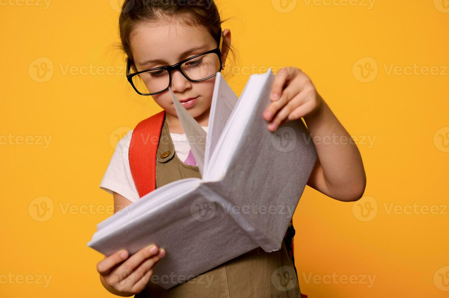 Smart elementary student kid girl wearing eyeglasses, learns reading alphabet, holding mockup book, isolated on orange photo